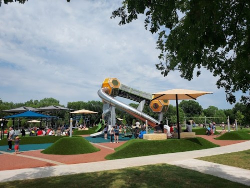 A vibrant playground with a large slide, children playing, and shaded areas under umbrellas on a sunny day.