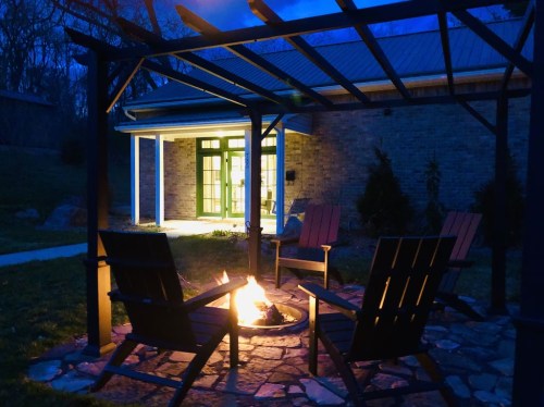 A cozy outdoor fire pit surrounded by wooden chairs, with a softly lit building in the background at dusk.
