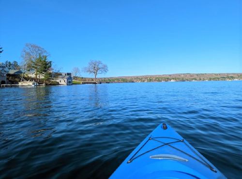 A view from a kayak on calm water, with a clear blue sky and distant shoreline featuring trees and houses.
