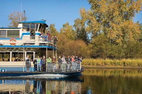 A tour boat with passengers cruising on a calm river, surrounded by trees with autumn foliage.