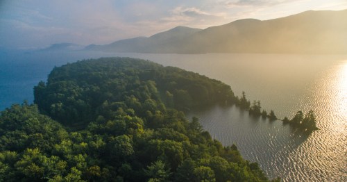 Aerial view of Lake George with a bit of fog, surrounded by green trees under a summer sky.