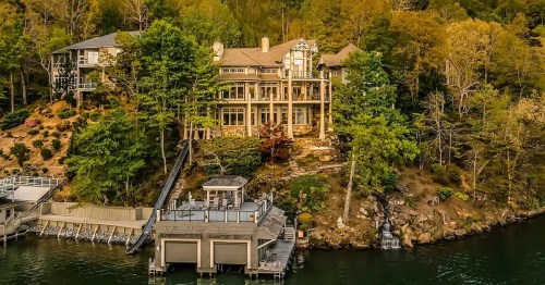 A large, elegant house surrounded by trees on a lakeside, with a dock and a small waterfall nearby.