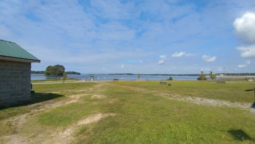 A serene lakeside view with blue skies, scattered clouds, and green grass in the foreground.