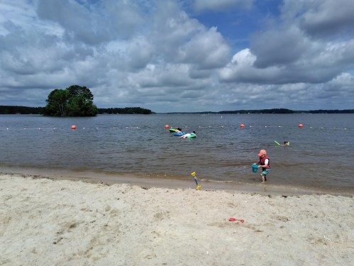 A sandy beach with a child playing near the water, colorful floats in the lake, and cloudy skies above.