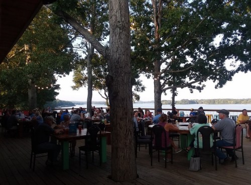 A lively outdoor dining area by a lake, filled with people enjoying meals under trees.