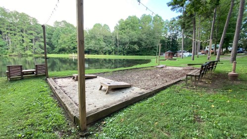 A serene outdoor area by a pond, featuring a sandy space for games, benches, and string lights among trees.