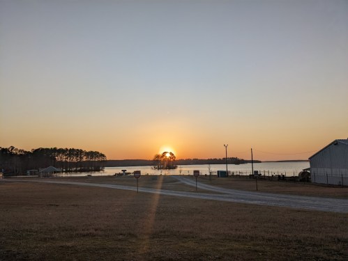 Sunset over a calm lake, with silhouettes of trees and a building in the foreground, casting a warm glow on the water.