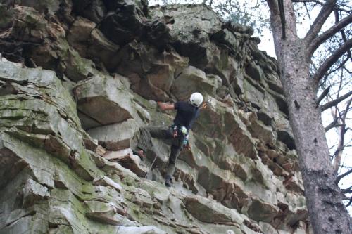 A climber ascends a rocky cliff, wearing a helmet and harness, surrounded by trees.
