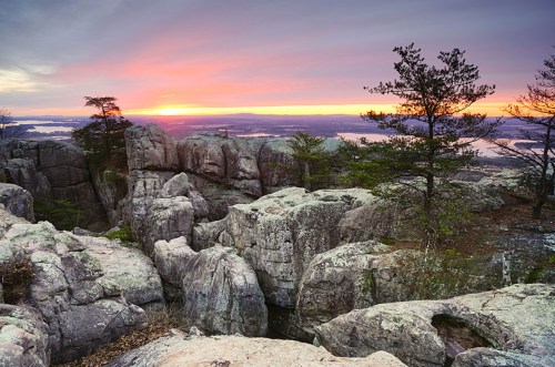 Sunset over rocky terrain with trees, casting warm colors across the sky and illuminating the landscape below.
