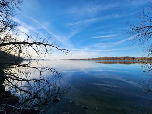 A serene lake scene with calm waters reflecting the sky, framed by bare tree branches.