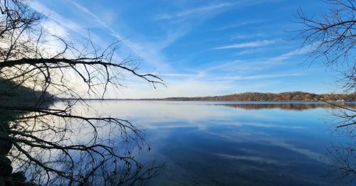 A serene lake scene with calm water reflecting a clear blue sky and distant hills, framed by bare tree branches.