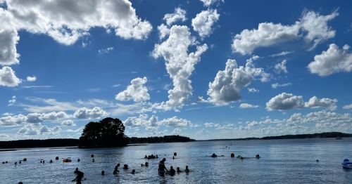 A sunny lake scene with people swimming and playing in the water, surrounded by fluffy clouds and a small island.