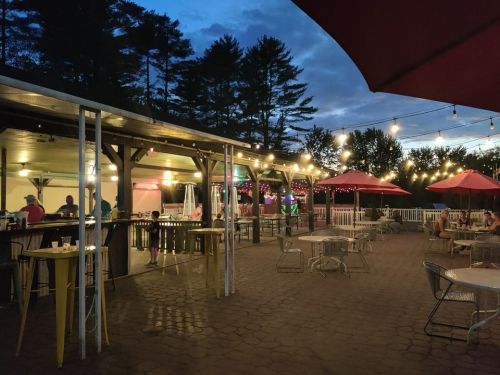 Outdoor dining area with string lights, tables, and umbrellas, set against a twilight sky and surrounded by trees.