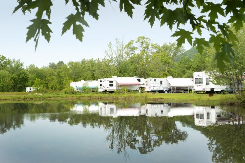 A row of RVs by a calm pond, surrounded by lush greenery and trees, reflecting in the water.