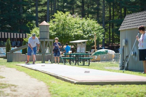 Two people play a game on a long outdoor court, surrounded by trees and picnic tables. A third person observes nearby.