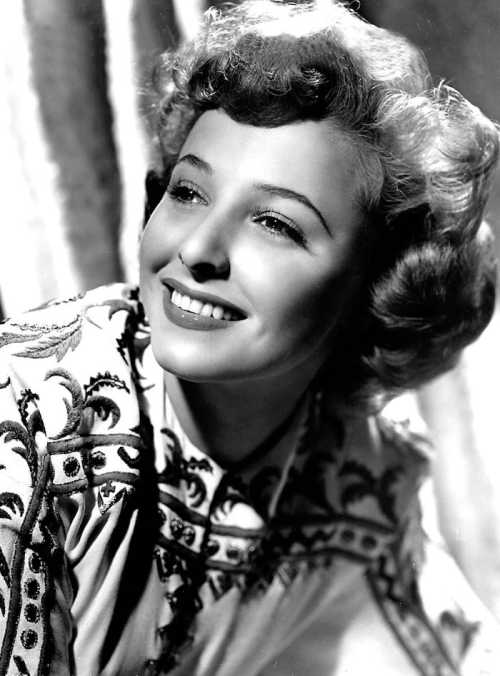 A vintage black-and-white portrait of a smiling woman with curly hair, wearing an embroidered blouse.