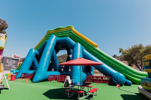 A large blue and green inflatable slide with a picnic table and umbrella nearby, set against a clear blue sky.