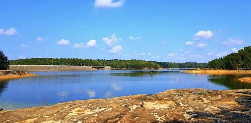 A serene lake surrounded by trees under a clear blue sky, with a dam visible in the distance.