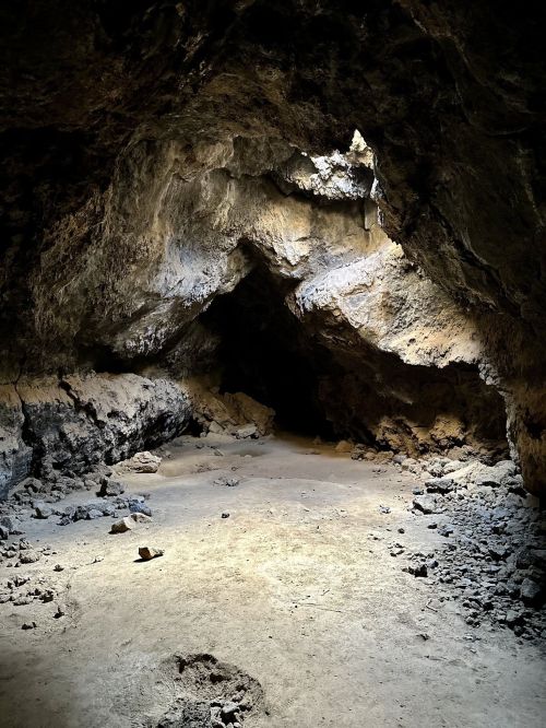 A dimly lit cave interior with rocky walls and a sandy floor, illuminated by light from an opening above.
