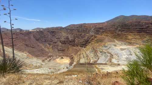 A vast open-pit mine with layered earth and rock formations under a clear blue sky, surrounded by mountains.