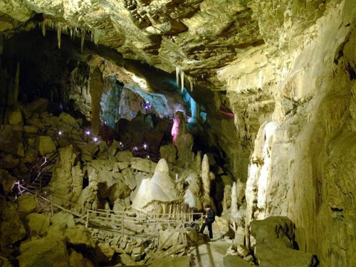 A dimly lit cave with rock formations, stalactites, and a wooden path, illuminated by colorful lights.