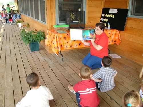 A woman reads a book to children sitting on the floor in a wooden outdoor setting.