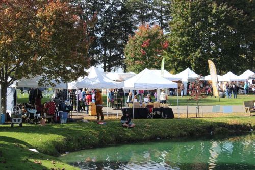 A vibrant outdoor market with white tents, vendors, and visitors near a pond surrounded by trees in autumn colors.
