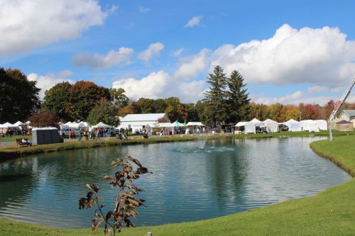 A serene pond surrounded by trees and tents at an outdoor event, with fluffy clouds in a blue sky.