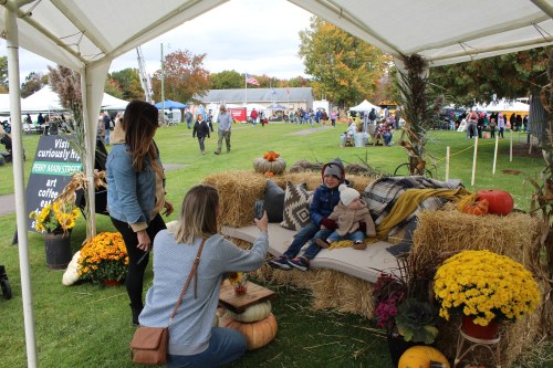 A family enjoys a fall festival, posing for a photo on a cozy couch decorated with pumpkins and flowers under a tent.