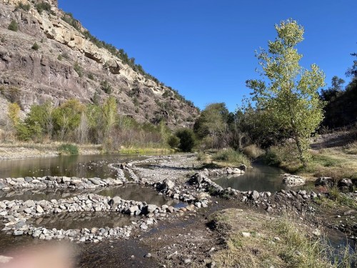 A serene landscape featuring a river, rocky shorelines, and trees against a backdrop of steep hills under a clear blue sky.