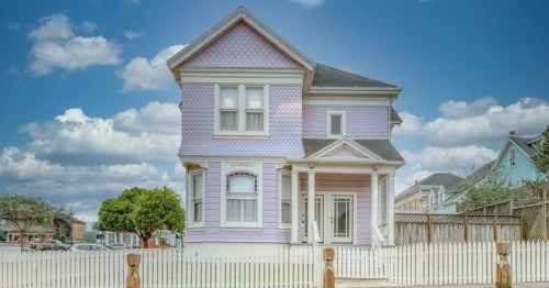 A charming purple Victorian house with a white picket fence under a blue sky with fluffy clouds.