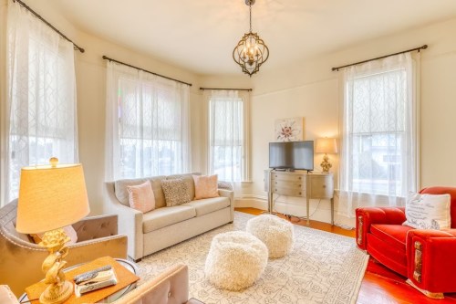 Cozy living room with a sofa, armchair, TV, and soft rug, featuring large windows and warm lighting.