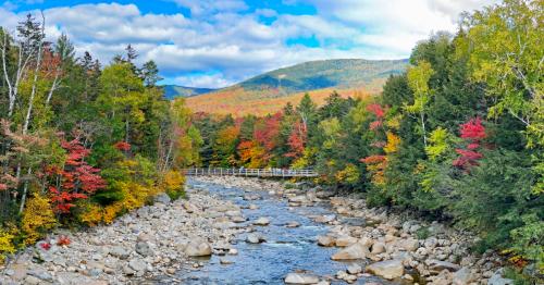 A scenic view of a river surrounded by colorful autumn foliage and mountains under a partly cloudy sky.