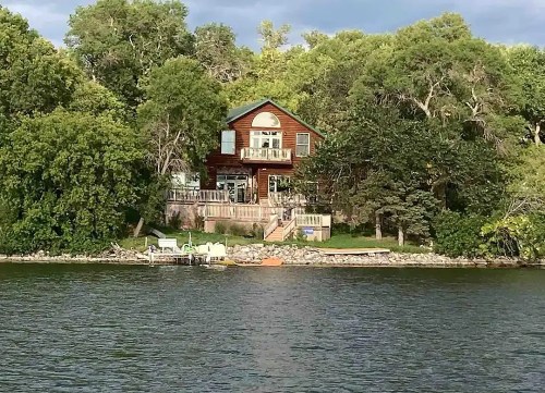 A wooden house surrounded by trees, located by a calm lake with a small dock and boats nearby.