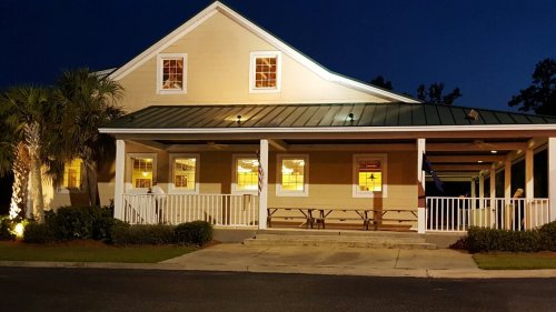A well-lit, two-story building with a green metal roof and porch, surrounded by palm trees at night.