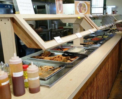 A buffet counter with various food trays, condiments in squeeze bottles, and labeled dishes in a casual dining setting.