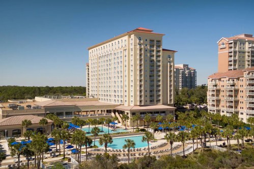A large hotel with a pool and palm trees, surrounded by other buildings under a clear blue sky.