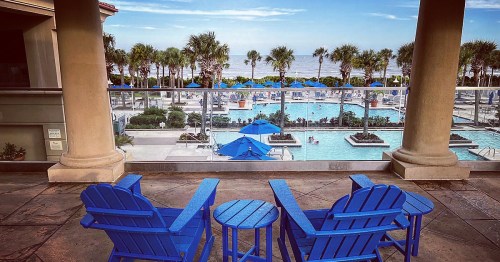 Two blue chairs and a small table overlook a pool area with palm trees and the ocean in the background.