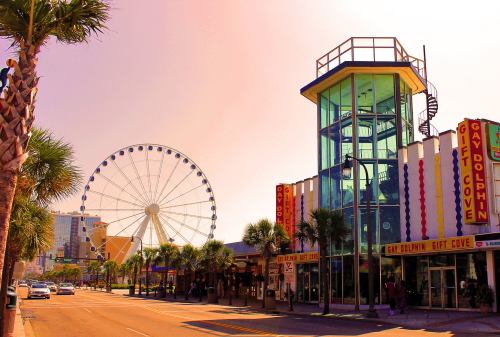 Colorful beachfront scene featuring a Ferris wheel, shops, and palm trees under a vibrant sky.