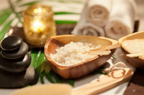 A serene spa setup with wooden bowls of salt, smooth stones, rolled towels, and a lit candle on a green leaf backdrop.