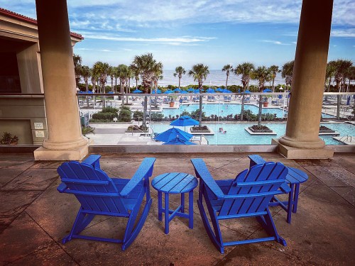 Two blue Adirondack chairs and a small table overlook a pool area with palm trees and the ocean in the background.