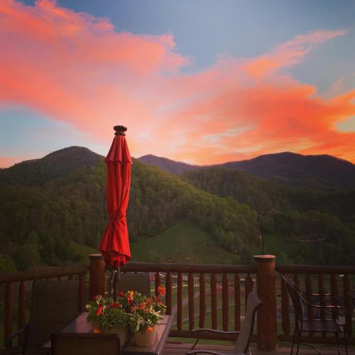 A vibrant sunset paints the sky in shades of pink and orange over green mountains, viewed from a deck with a red umbrella.