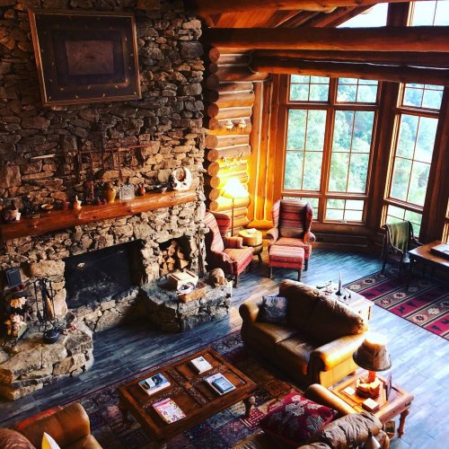 Cozy log cabin interior with a stone fireplace, large windows, and rustic furniture, creating a warm atmosphere.