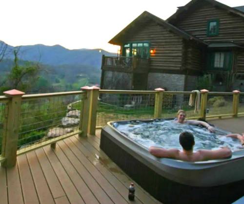 Two people relax in a hot tub on a deck, with a scenic mountain view and a log cabin in the background at sunset.