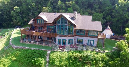 A large, rustic lodge with multiple windows, surrounded by greenery and a teepee in the background.