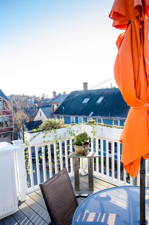 A sunny balcony with an orange umbrella, a table, and potted plants overlooking rooftops and a clear sky.