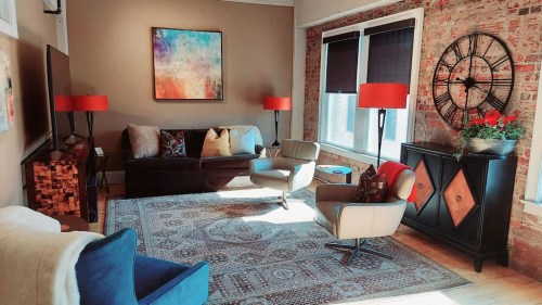 Cozy living room with a colorful abstract painting, red lamps, and a vintage clock on a brick wall.