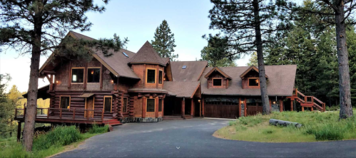 A large log cabin-style house surrounded by trees, with a winding driveway and a spacious lawn.