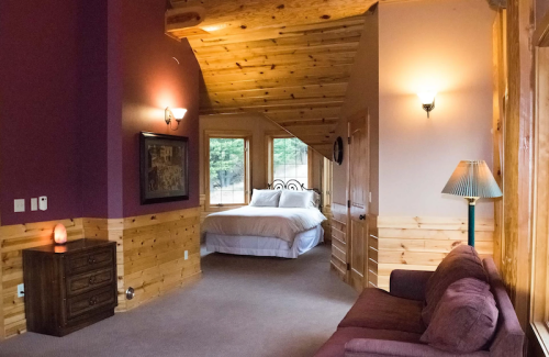 Cozy room with wooden accents, featuring a bed, sofa, and warm lighting. Large windows offer a view of the outdoors.