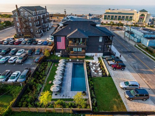 Aerial view of a coastal property with a pool, umbrellas, and an American flag, surrounded by parking and buildings.